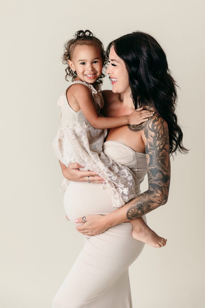 Pregnant woman wearing a beige maternity dress and holding her toddler daughter over pregnant belly. Mom is posing for her Maternity photoshoot orange county.
