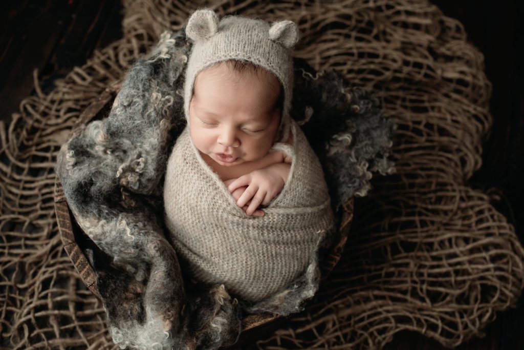 Newborn baby boy wrapped up in a brownish grey wrap and bear bonnet.