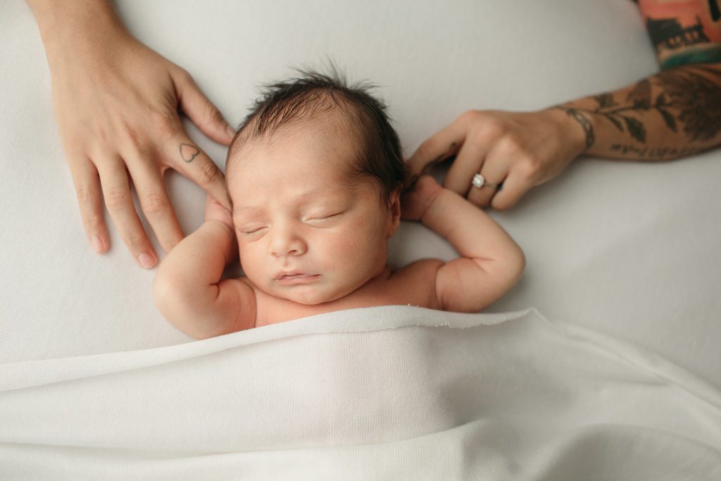 Image of a newborn baby boy lying in his back with his arms up and his mom holding his hands in hers.