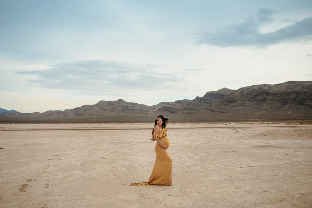 Pregnant woman standing sideways in a dry lake bed wearing a mustard off-shoulder maternity gown for her photoshoot.
